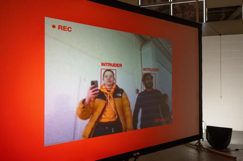 background is red. inside if a smaller recentage mimicing a video feed (you can see 2 people). Their faces are marked with a red rectange, with the word &quot;intruder&quot; and the left top corneer is a REC sign, bottom right corner is a timestamp