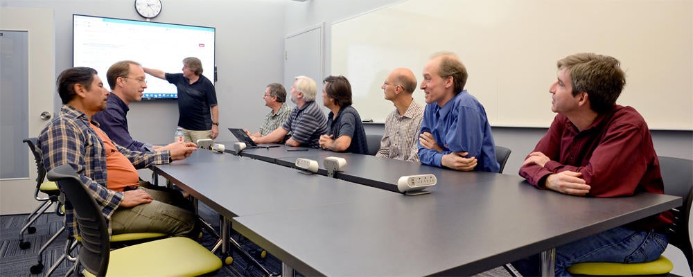 9 staff members sit around a meeting table, looking at a large display.