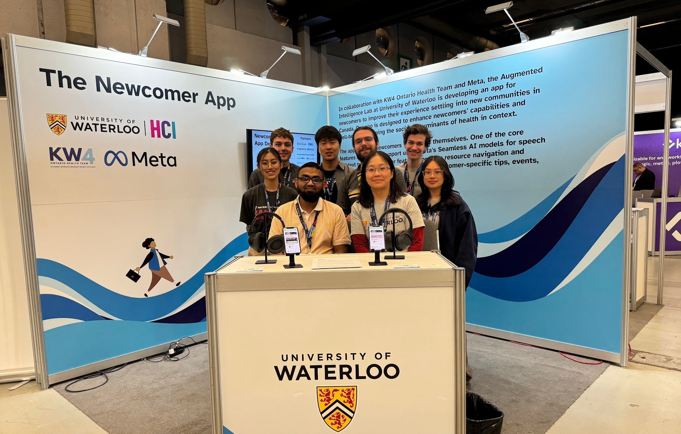 photo of the research team in front of their "The Newcomer App" booth at a conference. The table has 2 phones with 2 headphones