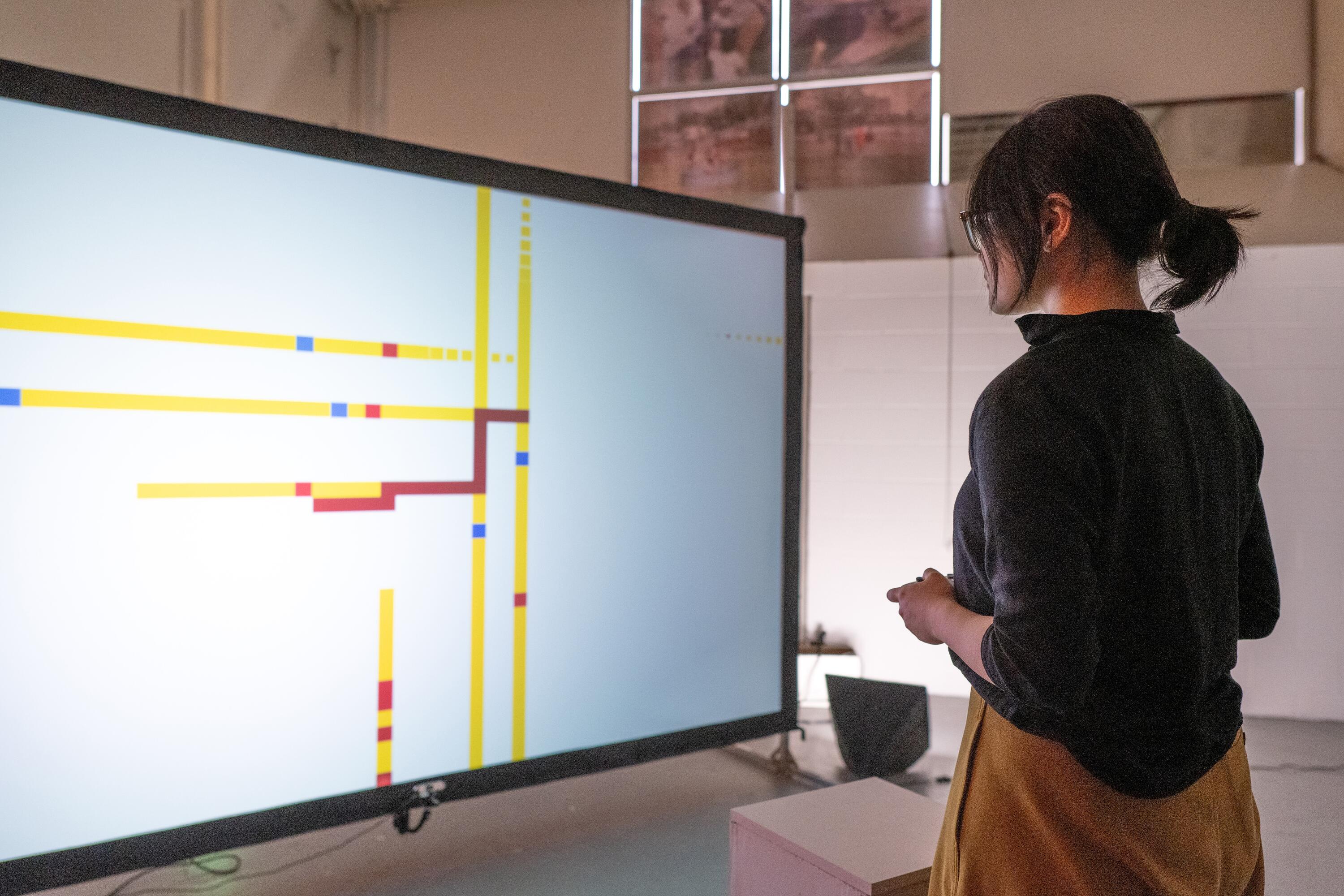 Student playing a revised verison of snake: standing in front of a screen holding a controller. back is facing the camera