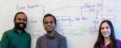 Three people (two men on the left and one woman on the right) stand in front of a whiteboard filled with a flowchart; left wears a green sweater, middle a gray dotted shirt, right a purple top.
