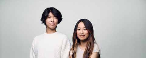 An Asian male donning a white shirt and grey slacks standing next to an Asian female (sitting on a stool) wearing a light colour shirt and brown pants. They pose against a grey background