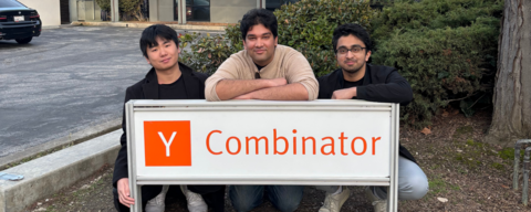 Co-founders crouching in front of the Y Combinator sign (white sign. The "Y" is in an orange block, with white font. Combinator is in orange font). Photo taken outdoors in front of a white building