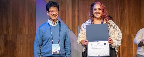 An Asian man in blue standing next to a Middle Eastern woman wearing a black dress and a white lacey jacket. She is holding her award