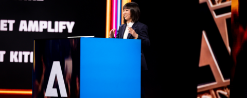 An asian woman speaking at a podium with a black and colourful neon lights background