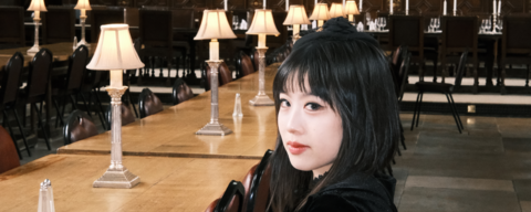 A person in a black velvet dress and gloves sits at a long wooden table in a historic dining hall with decorative wall panels and lamps. A large painting hangs on the wall behind them.