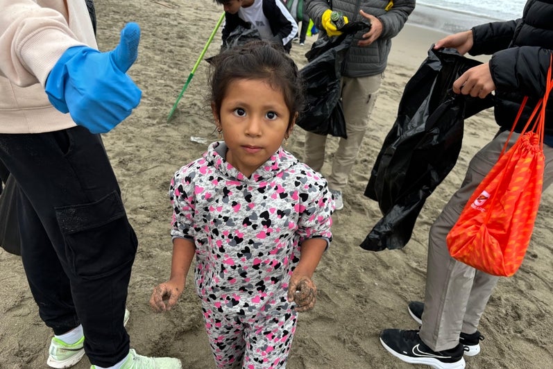 A close-up of the people cleaning a beach (holding trash bags). You can see someone's hand donning a blue cleaning glove and giving a thumb-ups (face not visible). In the middle is a young girl facing the camera