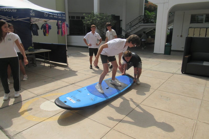 A young boy wearing a school uniform and standing on a surfboard while being on pavement. A young man is crouching down at the side of the surboard to hold it down.  