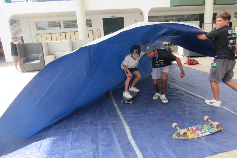A young boy on a skateboard with someone holding him. Another man is holding a blue-coloured cloth on top of the boy and his partner, emulating a wave