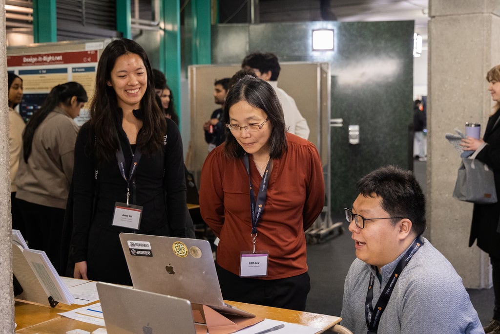 A group of people is gathered at a table with laptops. Two individuals stand, while one person is seated, looking at the screen. They are attending a poster session.