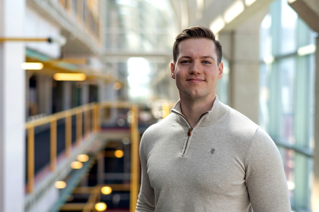 A man wearing a light gray sweater stands in a modern indoor space with large windows and a blurred background.