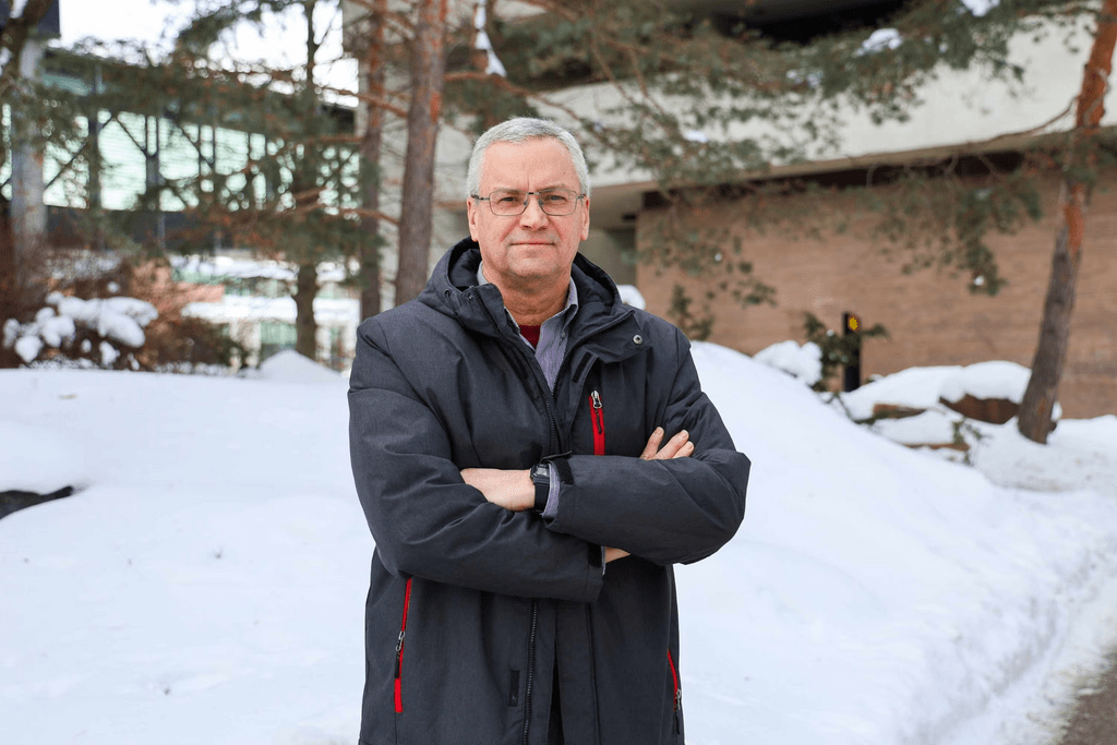 A man donning a black winter jacket posing in front of snow and brown buildings