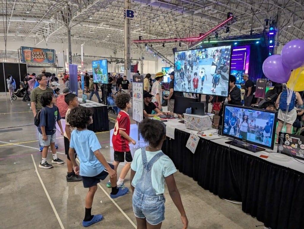 Group of children standing in a straight horizontal line and playing a soccer-like game. They are standing in front of a large tv screen and a laptop screen, which depict a VR game where the user is kicking a soccer ball 
