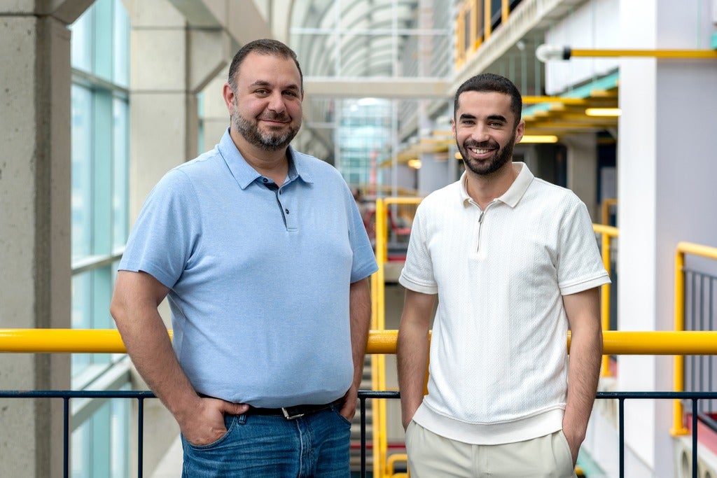 Two men (the left person wearing a baby blue collared shirt with jeans and the right person wearing a white collared shirt with light grey pants) posing in front of a yellow rail