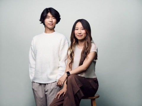 An Asian male donning a white shirt and grey slacks standing next to an Asian female (sitting on a stool) wearing a light colour shirt and brown pants. They pose against a grey background