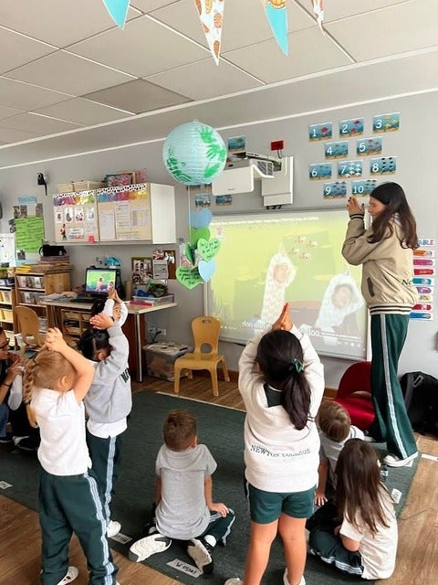 A group of kindergartners holding their hands above their head (a clap position/looks like a triangle) in front of a projector (screen is playing baby shark). A woman wearing green pants and a grey-brown hoodie is leading the session
