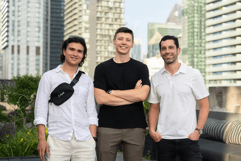 Three men (left man is wearing a white button-up shirt with a cross-bag, middle man is crossing his arms and wearing black shirt with grey pants, and the right man is wearing a white polo shirt) posing in front of skyscrapers