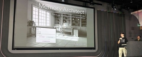 Stage presentation with a large screen titled “Memory Architect”; grayscale slide shows a room with shelves and a desk. Two presenters stand to the right, one speaking into a microphone.