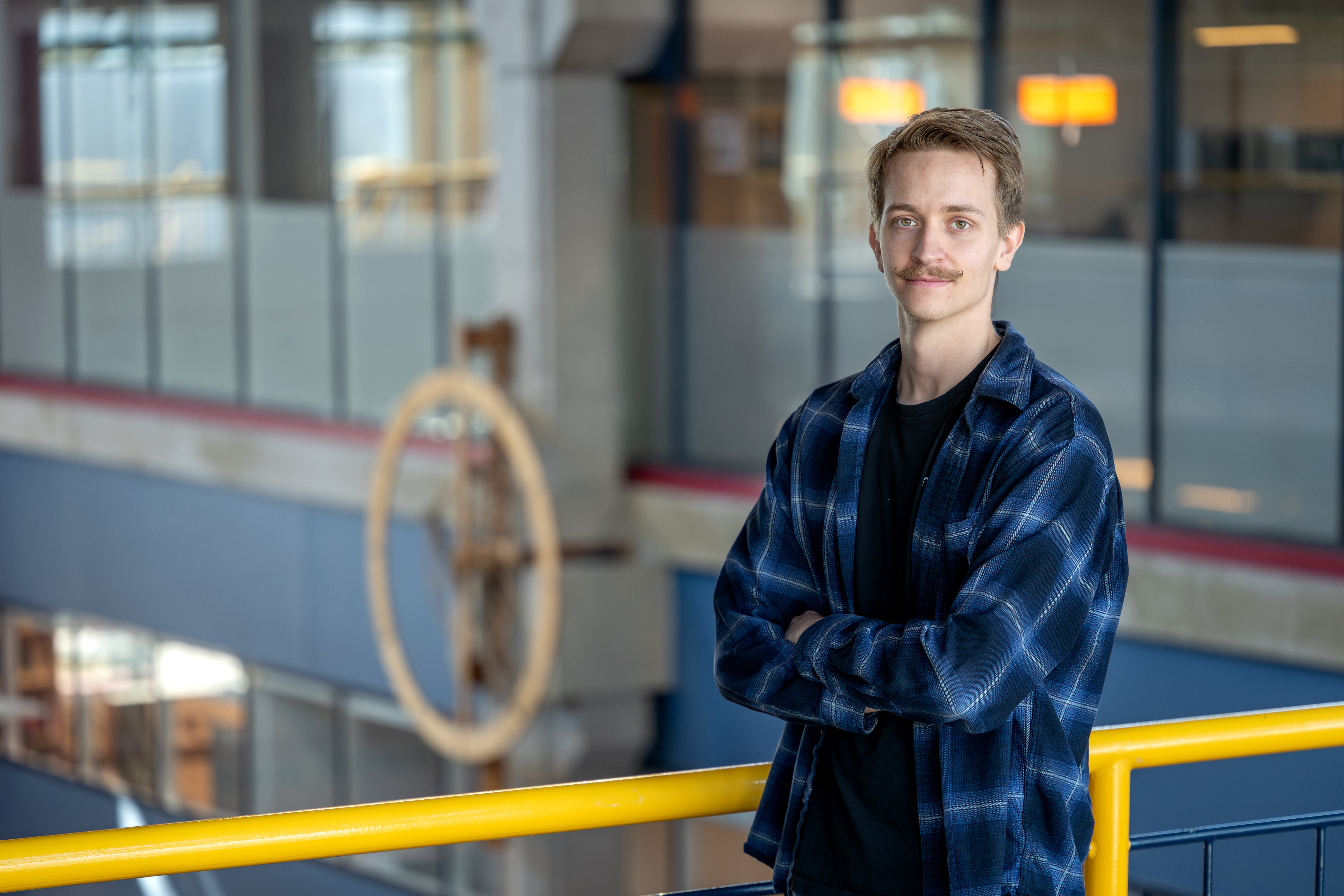 a blond wearing a dark blue plaid shirt on top of a black shirt. he poses aganist a yellow rail. There is a wooden clock in the background