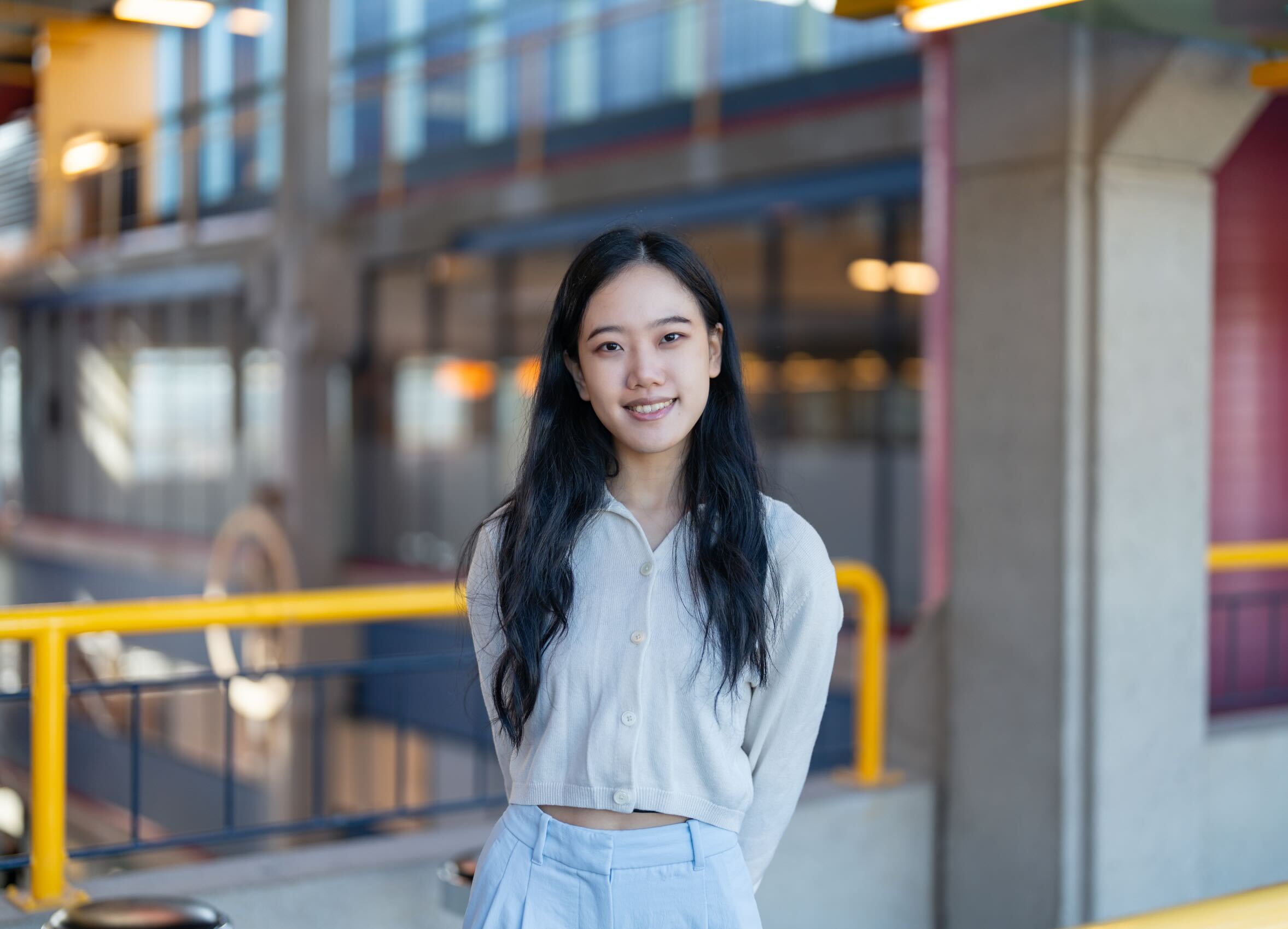 An Asian woman wearing a collared grey shirt and blue jeans posing in front of the yellow rail in Davis Centre