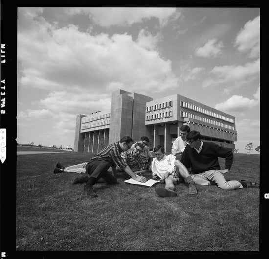 Black and white photo photo of 6 men studying and sitting on grass. In the middle of the men there is a book, where one of the men is writing in it. In the backdrop you see the MC building.