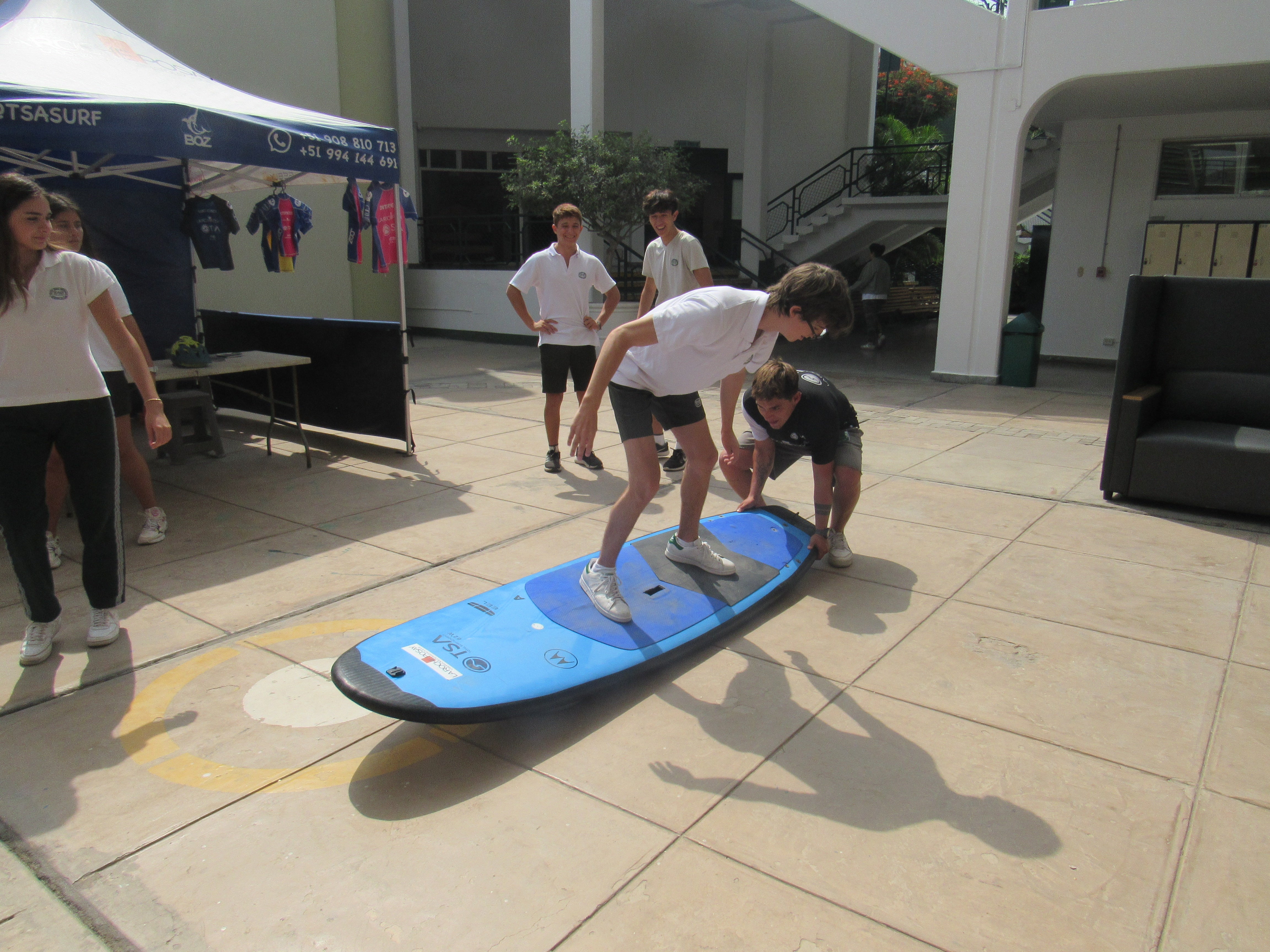 A young boy wearing a school uniform and standing on a surfboard while being on pavement. A young man is crouching down at the side of the surboard to hold it down.  