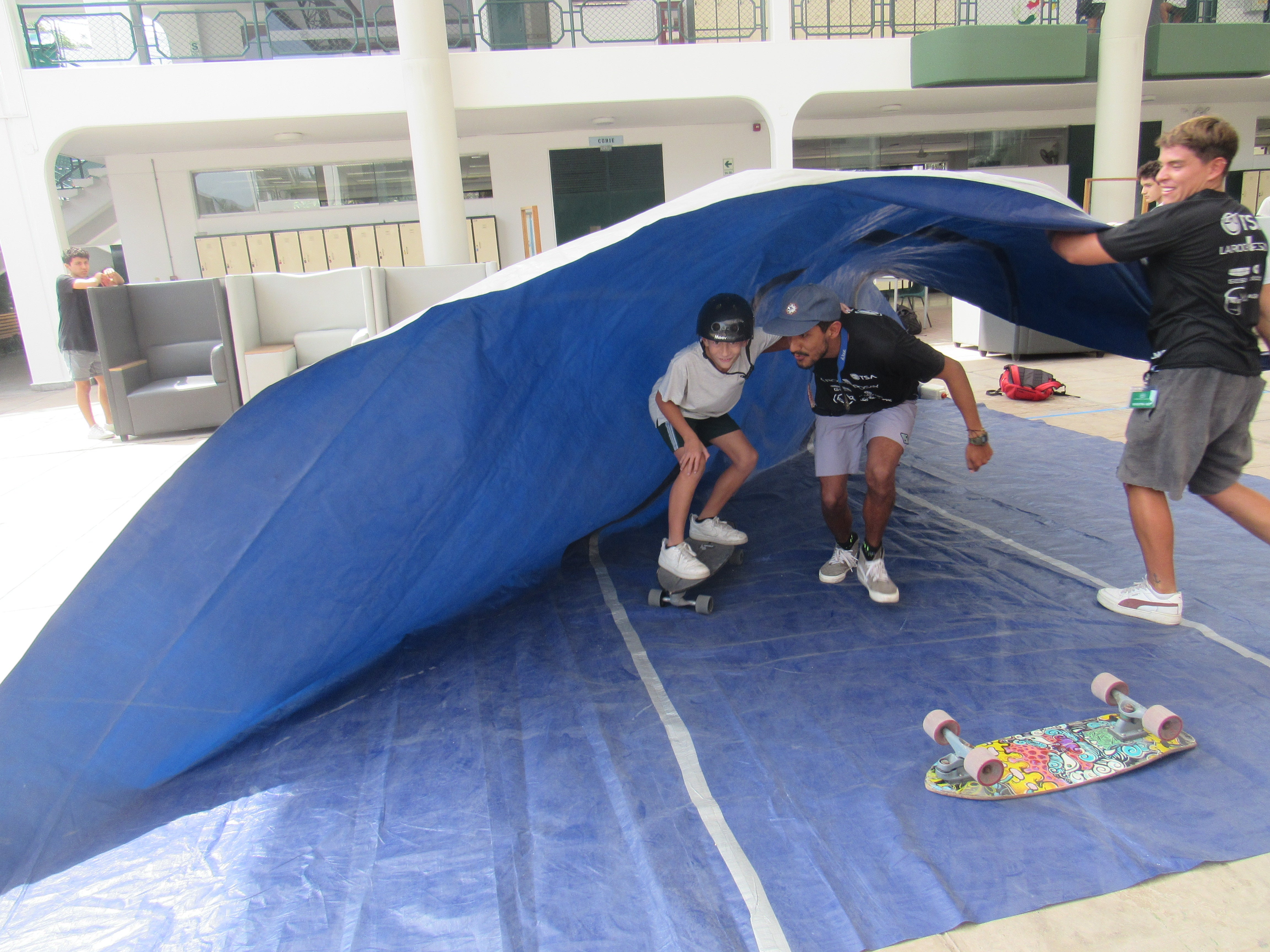 A young boy on a skateboard with someone holding him. Another man is holding a blue-coloured cloth on top of the boy and his partner, emulating a wave