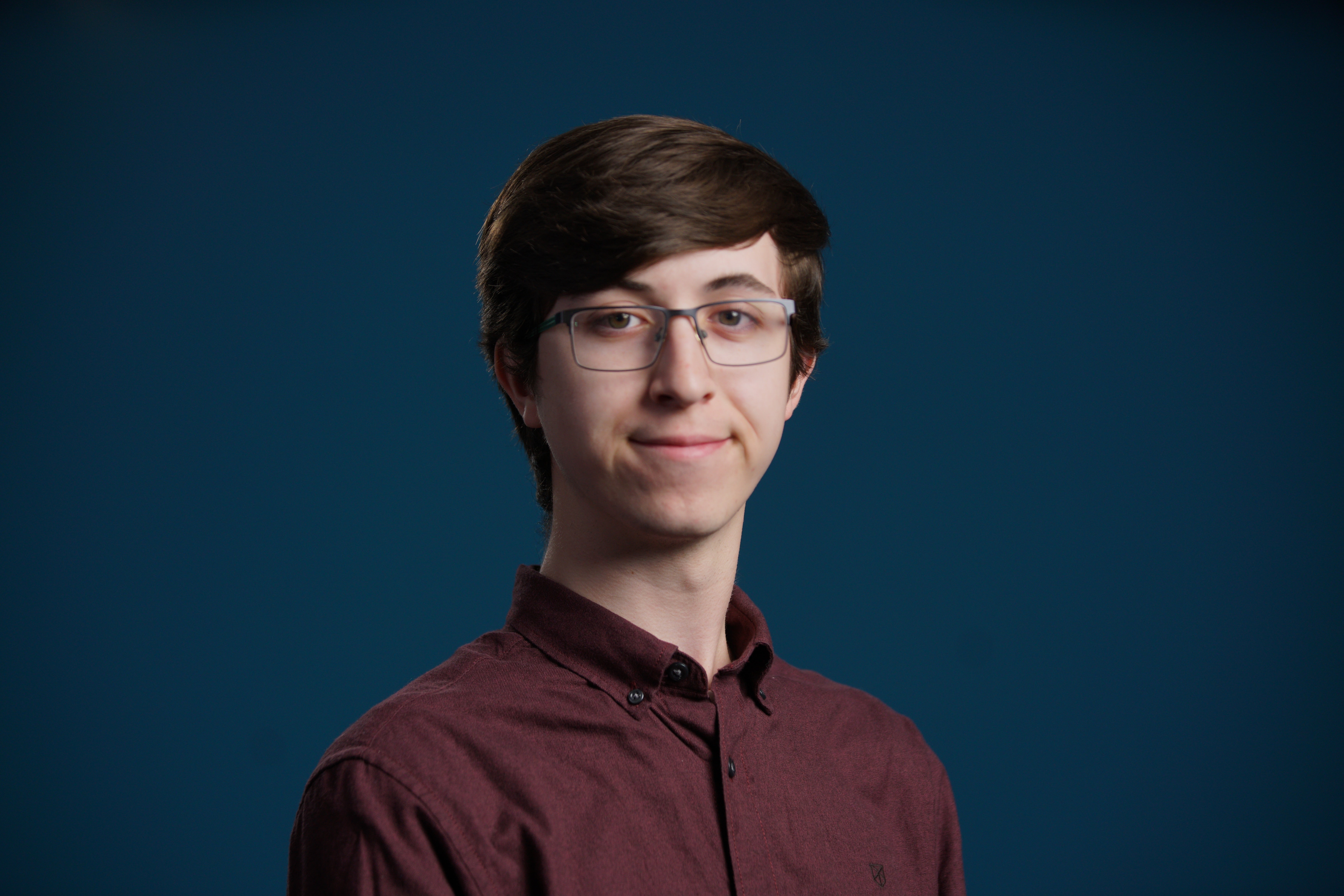 A Caucasian man with in a maroon/burgundy collared shirt posing aganist a blue background. He has brown hair and wears glasses