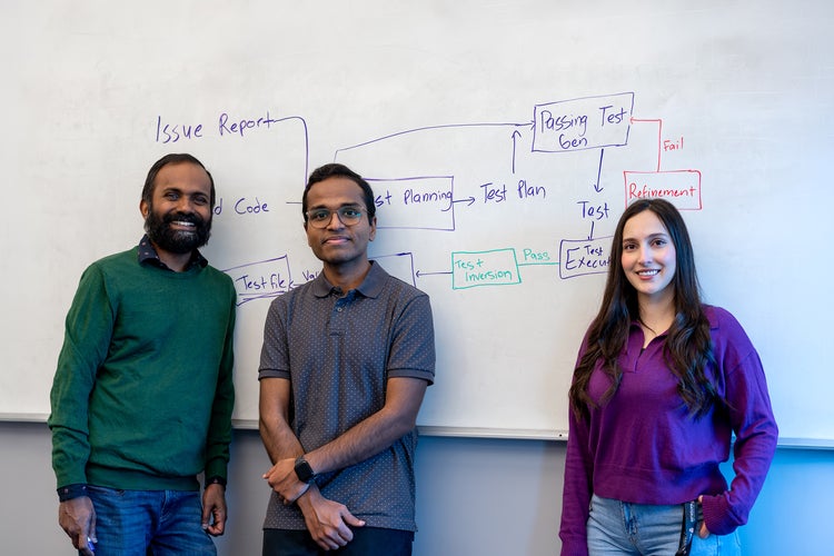 Three people (two men on the left and one woman on the right) stand in front of a whiteboard filled with a flowchart; left wears a green sweater, middle a gray dotted shirt, right a purple top.