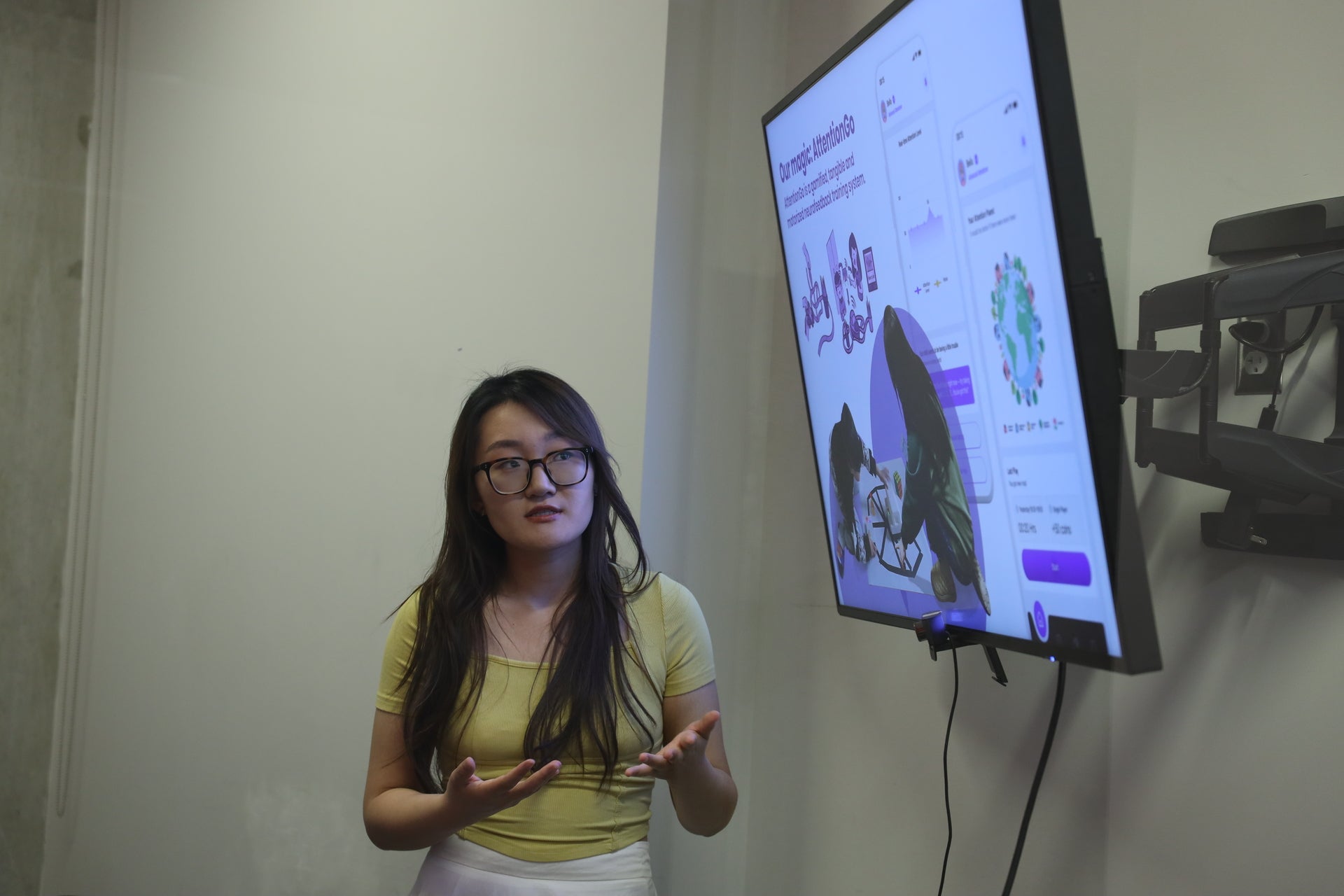 An Asian woman wearing a yellow shirt presenting her research pitch to an audience (not shown). At her side is a large tv displaying her presentation