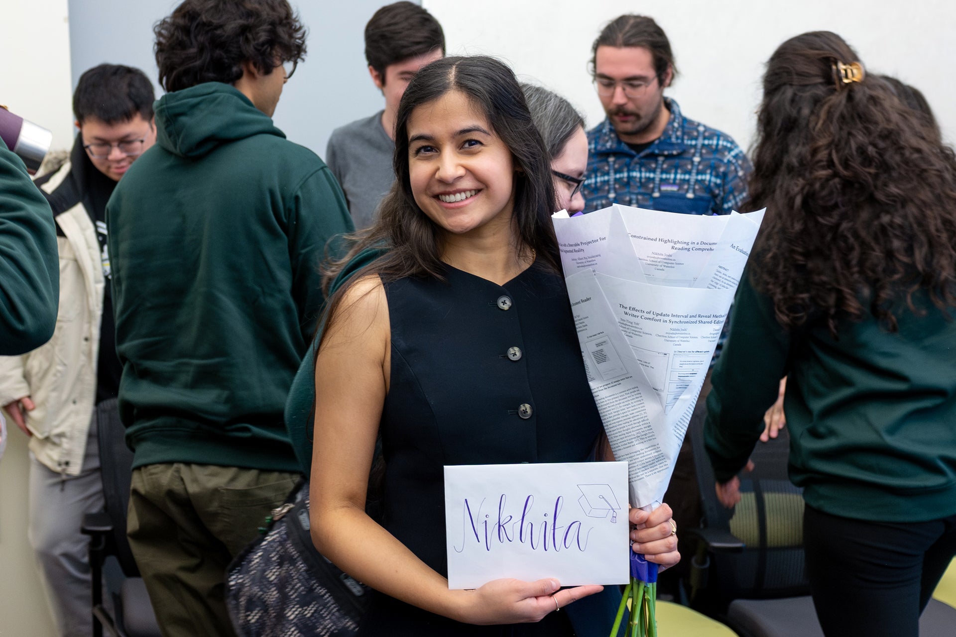Nikhita donning a navy-blue dress holding a bouquet (which is composed of her research papers) and an envelope that says "Nikhita"