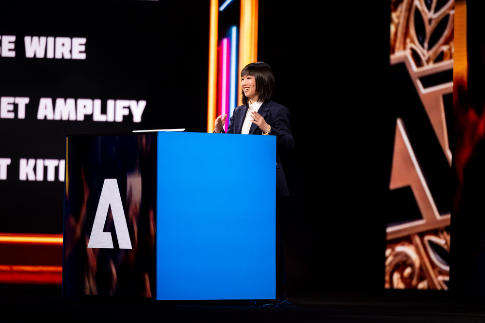An asian woman speaking at a podium with a black and colourful neon lights background